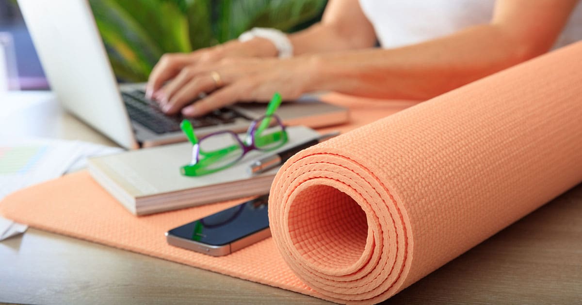 Yoga mat half rolled out with cell phone, notebook and glasses on top with someone using a laptop in the background